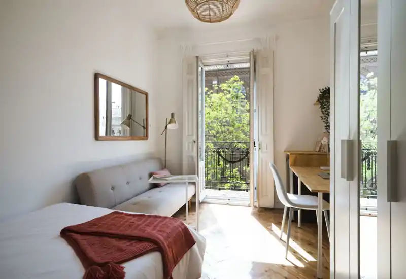Sunny, cozy bedroom with a comfortable bed, a compact sofa and a work desk opening onto a small balcony with greenery — bright, stylish and functional for short stays.