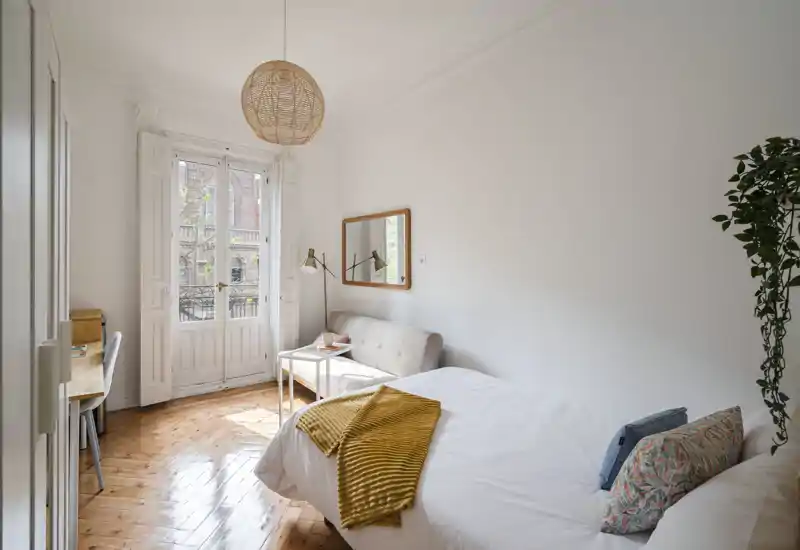 Bright, stylish bedroom with a comfortable double bed, cosy throw and cushions, a small sofa and desk by large French doors letting in abundant natural light.