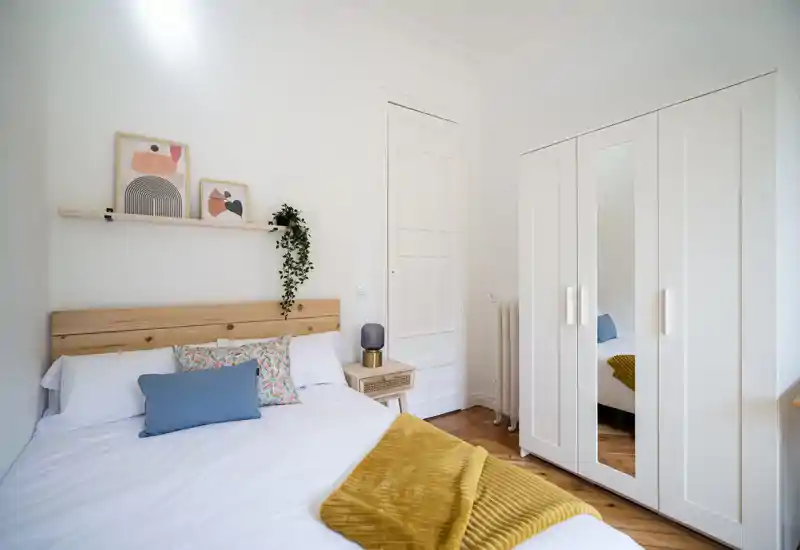 Bright, minimal bedroom with a wooden headboard, patterned cushions and a mustard throw; a white wardrobe with mirrored door expands the space.