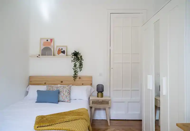 Bright, cozy bedroom with a wooden headboard, patterned cushions and a mustard throw. Clean white walls, a bedside table with lamp and a wardrobe with mirrored doors create a fresh, airy feel.