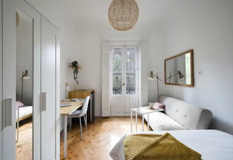 Bright, stylish bedroom with a comfortable bed in the foreground, a small sofa and a desk by a tall window that brings abundant natural light—ideal for relaxing and working.