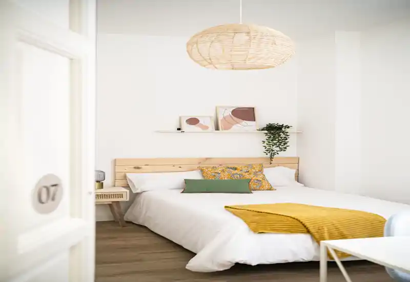 Bright, minimal bedroom with a wooden headboard, patterned cushions and a mustard throw. Clean white linens, a hanging woven pendant and a small shelf with art create a cozy, modern feel ideal for rental listings.