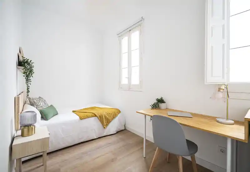 Chambre simple lumineuse et minimaliste avec lit confortable, table de chevet en bois et bureau près de la fenêtre — idéale pour un étudiant ou une personne seule.