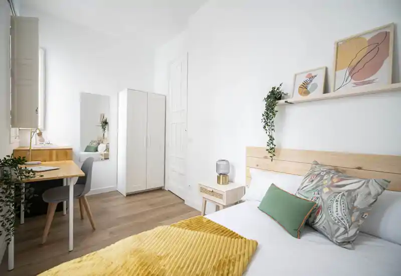 Chambre lumineuse et cosy avec tête de lit en bois, coussins décoratifs et plaid moutarde. Un petit bureau et une armoire rendent la pièce fonctionnelle pour le travail et le rangement.