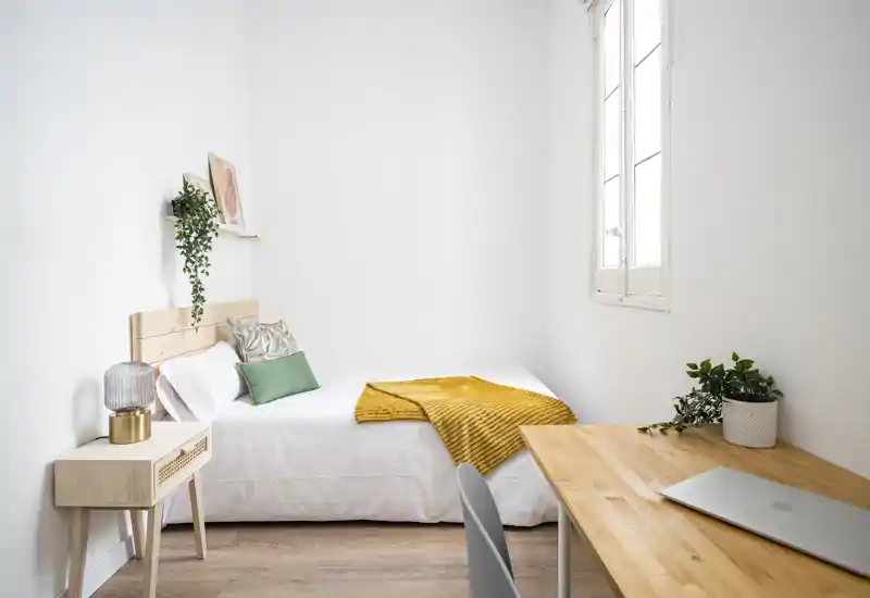 Chambre lumineuse et minimaliste avec un lit bien fait, tête de lit en bois, coussins verts et un plaid moutarde; un bureau en bois près de la fenêtre et des plantes ajoutent une ambiance moderne et apaisante.