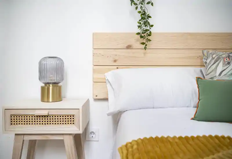 Coin chambre lumineux et élégant avec tête de lit en bois, linge de lit blanc, coussins décoratifs et table de chevet avec lampe en verre — atmosphère calme et accueillante.