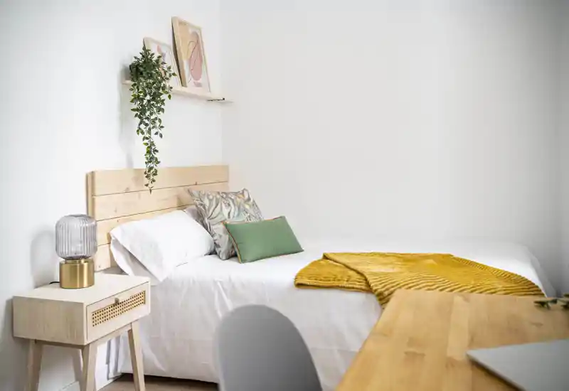 Chambre lumineuse et minimaliste avec tête de lit en bois, linge de lit blanc, coussins décoratifs et un plaid moutarde ; table de chevet avec lampe et petite plante sur une étagère pour une ambiance scandinave chaleureuse.