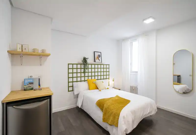Bright, modern single bedroom with a neatly made bed, cheerful yellow accents and minimal, stylish decor. Natural light from a window and clean white walls create an airy, welcoming atmosphere.