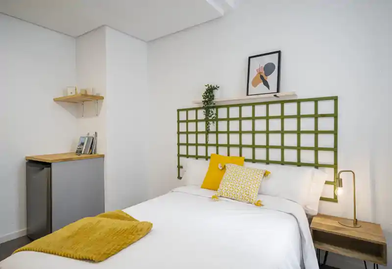 Chambre lumineuse et accueillante avec un lit double bien fait, textiles jaunes en accent, table de chevet avec lampe et décor mural minimaliste.