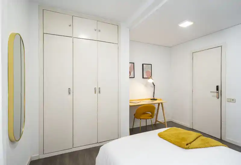 Chambre moderne et lumineuse avec lit bien fait, dressing intégré et un petit bureau. Murs blancs et accents jaunes coordonnés créent une atmosphère fraîche et accueillante.