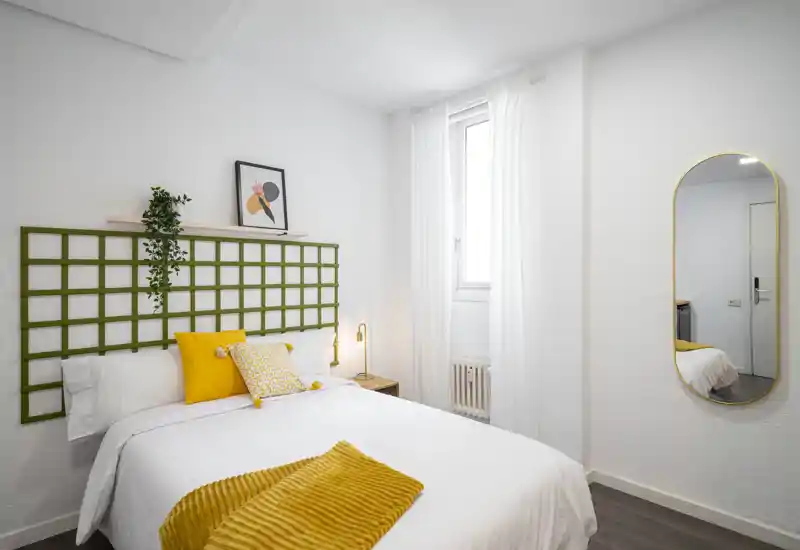 Bright, modern bedroom with a neatly made double bed, warm yellow accents and natural light from a window.
