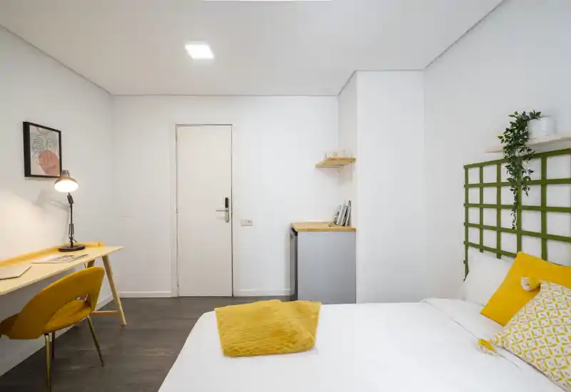 Chambre lumineuse et moderne avec un lit bien fait, des accents jaunes chaleureux et un petit espace de travail. Idéale pour une personne seule ou un couple recherchant une pièce cosy et bien éclairée.
