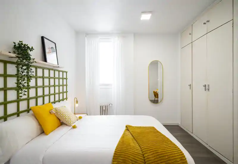 Chambre lumineuse et moderne avec un lit double bien fait, accents moutarde chaleureux et placard intégré. La pièce paraît aérée grâce à la lumière naturelle et à une décoration contemporaine et épurée.