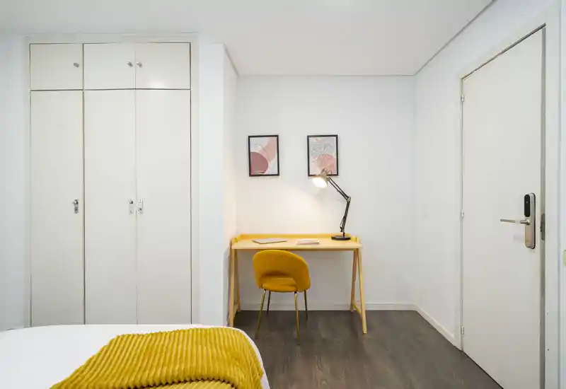 Coin chambre lumineux et soigné : lit visible au premier plan, bureau et chaise jaunes contre le mur et une lampe apportant une lumière chaude.
