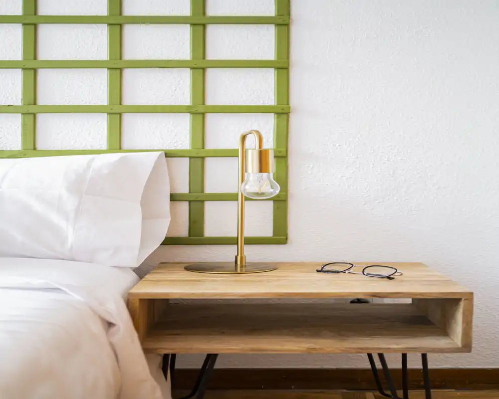 Coin de chambre lumineux et minimaliste avec draps blancs, table de chevet en bois et une lampe en laiton moderne devant une tête de lit en treillis vert — ambiance calme et bien éclairée.