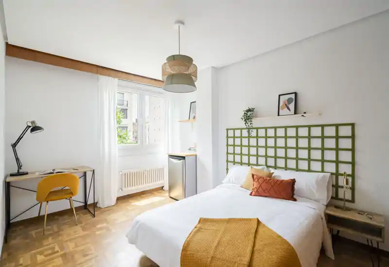 Chambre lumineuse et soignée avec lit double blanc, touches moutarde et un petit bureau près de la fenêtre — parfaite pour un couple ou un séjour individuel.