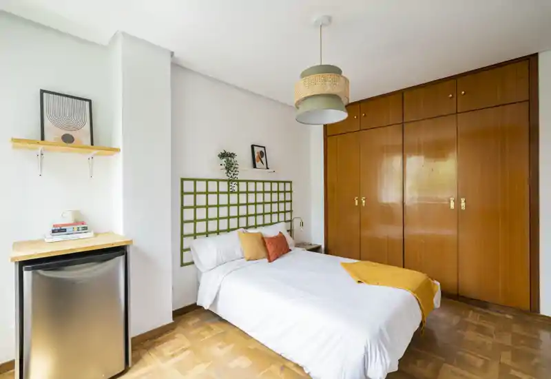 Chambre lumineuse et bien aménagée avec un lit double, une grande armoire en bois et des coussins décoratifs. La lumière naturelle et les touches chaleureuses rendent la pièce accueillante.
