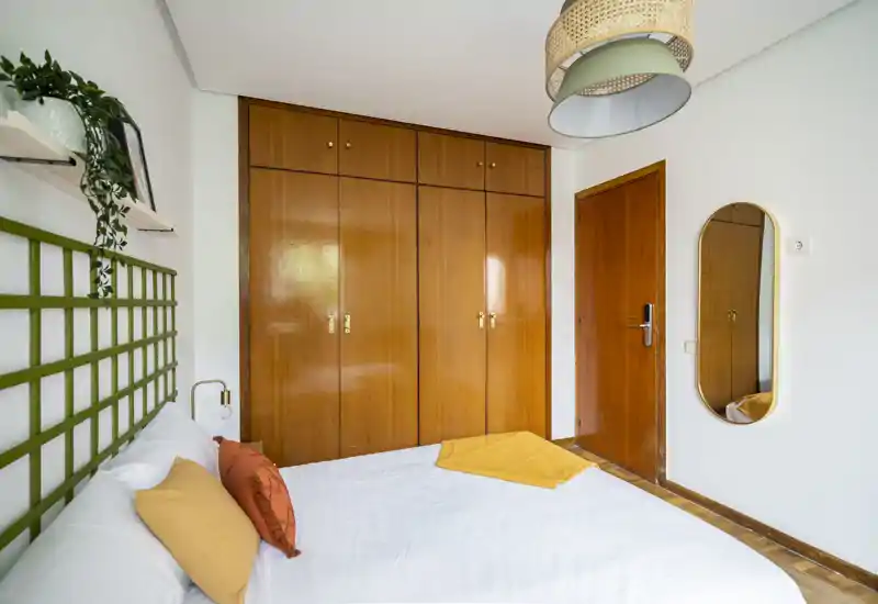 Chambre lumineuse et élégante avec grand placard mural en bois, coussins décoratifs et touches chaleureuses ; literie blanche propre et suspension qui créent une ambiance accueillante.
