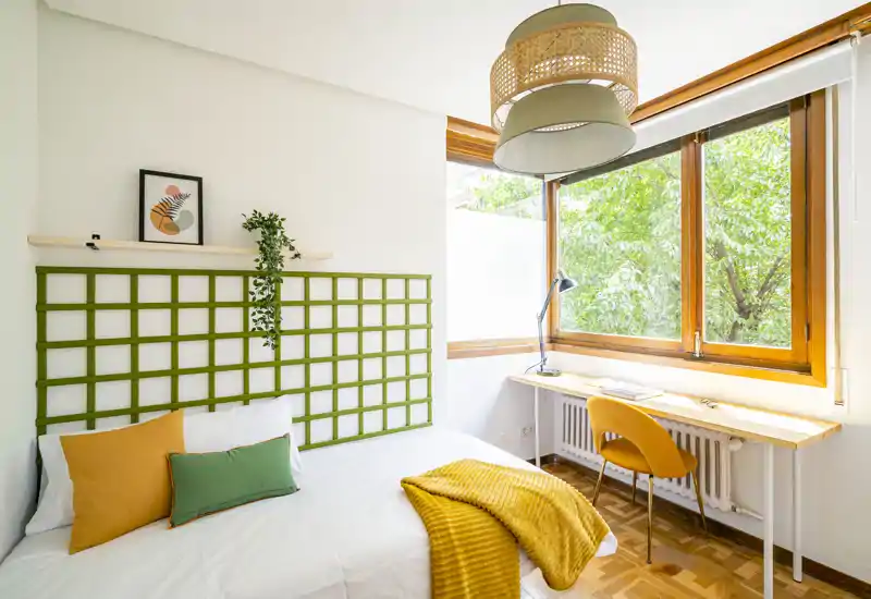 Chambre lumineuse et chaleureuse avec un lit bien fait, des coussins colorés et un plaid douillet ; une fenêtre à cadre bois apporte une belle lumière naturelle et un petit bureau offre un coin travail.