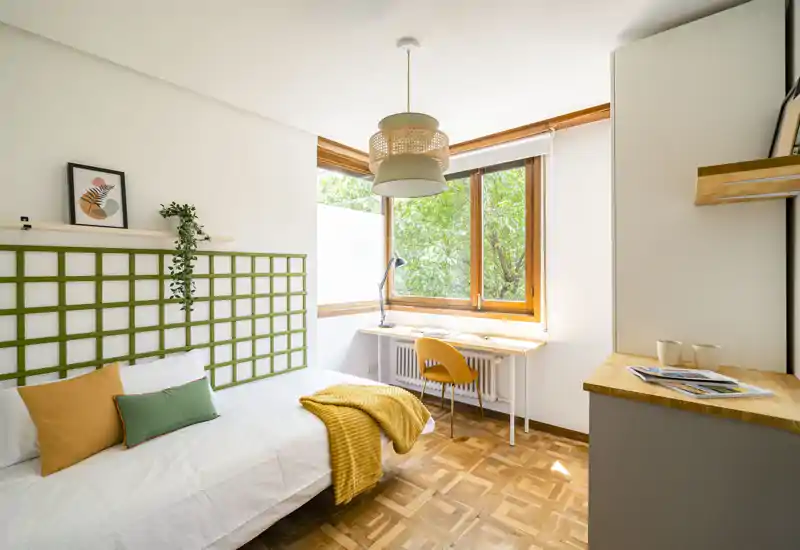 Chambre simple lumineuse et soignée avec un lit confortable, coussins décoratifs et un plaid jaune chaleureux. Grande fenêtre apportant beaucoup de lumière naturelle et un petit bureau près de la fenêtre idéal pour étudier.