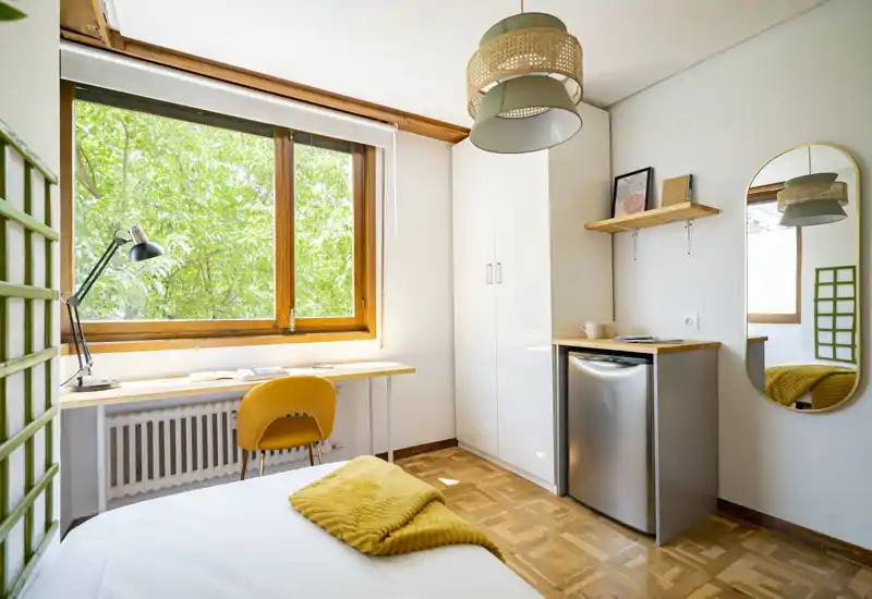 Chambre simple lumineuse et bien décorée avec grande fenêtre, sol en bois, bureau compact et armoire. Touches jaunes chaleureuses (plaid et chaise) et lumière naturelle douce pour un séjour confortable.