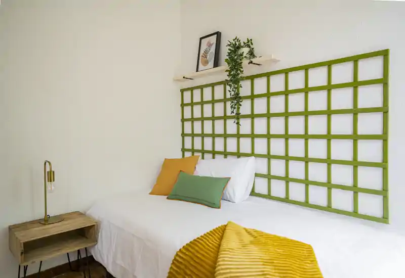 Chambre simple lumineuse et stylée avec lit blanc, coussins colorés et plaid jaune texturé ; table de chevet en bois et treillis mural décoratif pour une ambiance chaleureuse.