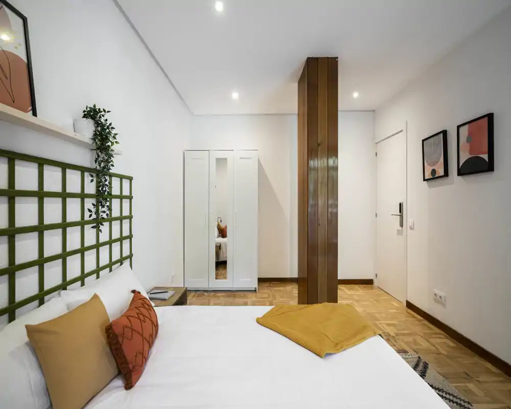 Camera da letto moderna e luminosa con letto ordinato, cuscini decorativi e un plaid giallo. Pavimento in parquet e arredo minimale creano un ambiente accogliente e tranquillo, perfetto per annunci.
