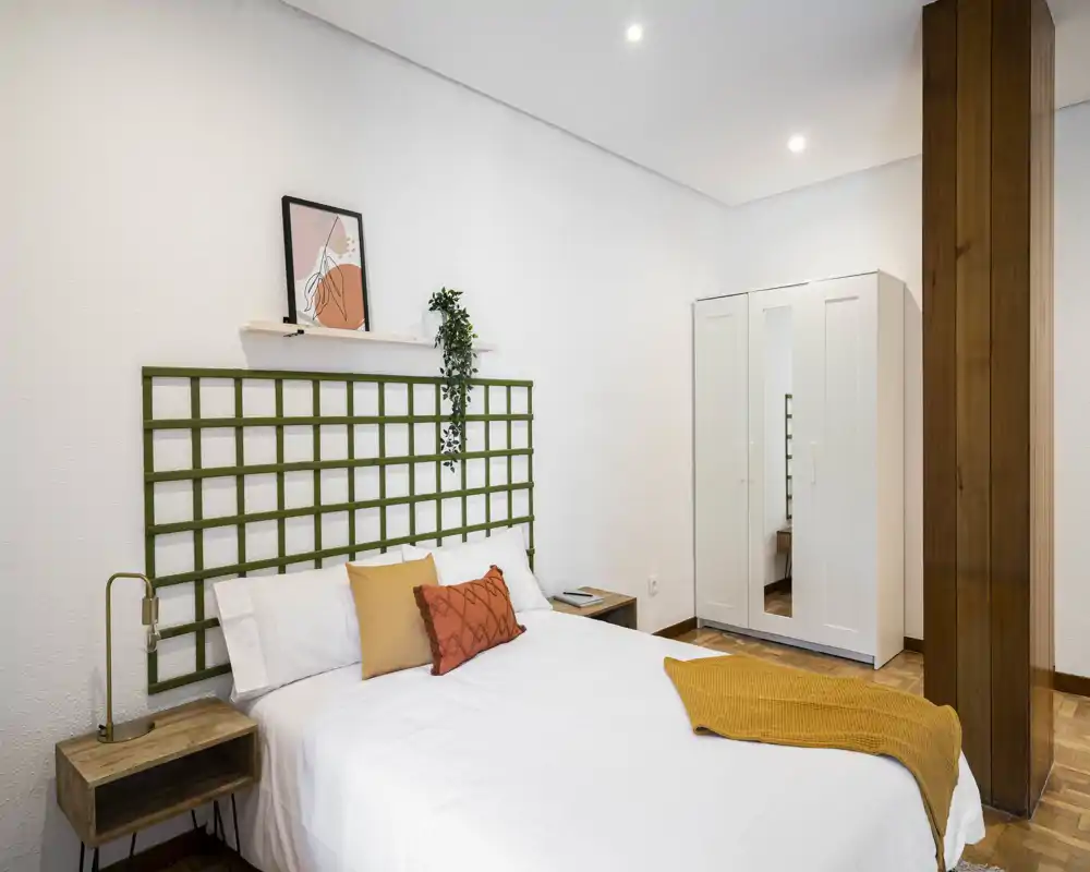 Camera da letto luminosa e moderna con letto matrimoniale, cuscini decorativi e plaid senape; pareti bianche, comodini e un armadio con specchio per pratico spazio di stoccaggio.