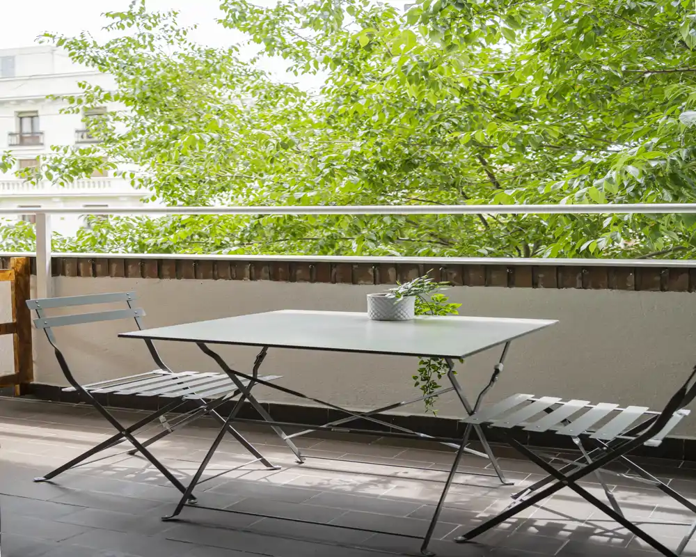 Balcone soleggiato con un semplice tavolo in metallo e due sedie pieghevoli, una piccola pianta sul tavolo e alberi frondosi oltre la ringhiera — ideale per il caffè mattutino.
