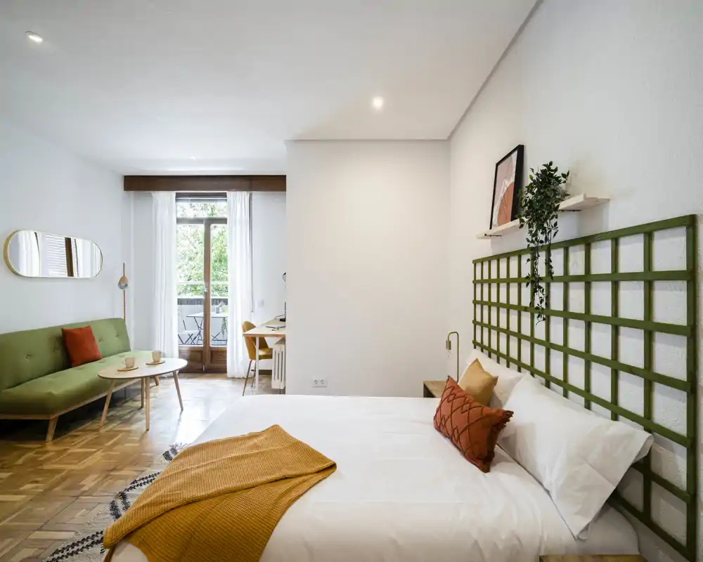 Camera da letto luminosa e di design con letto matrimoniale comodo, tessili caldi e un piccolo angolo salotto vicino al balcone. Grande finestra/porta che offre molta luce naturale e vista sul verde.