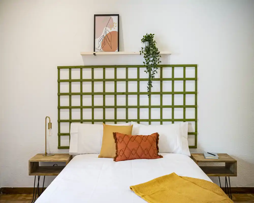 Camera da letto luminosa e ben arredata con un grande letto in lenzuola bianche, cuscini dalle tonalità calde e un plaid giallo senape; testiera decorativa, comodini e una pianta sospesa rendono l'ambiente accogliente e moderno.