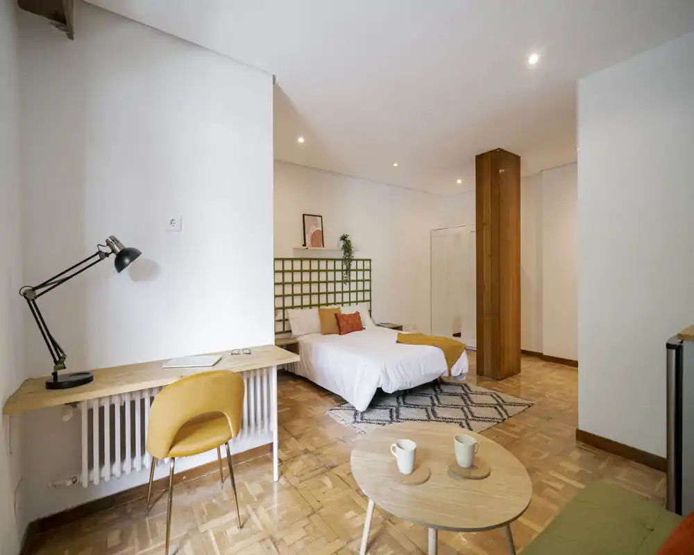 Camera da letto moderna e luminosa con letto matrimoniale, cuscini decorativi e tappeto a motivo; una scrivania in legno con sedia crea un angolo studio accogliente e il parquet caldo unisce l'ambiente.