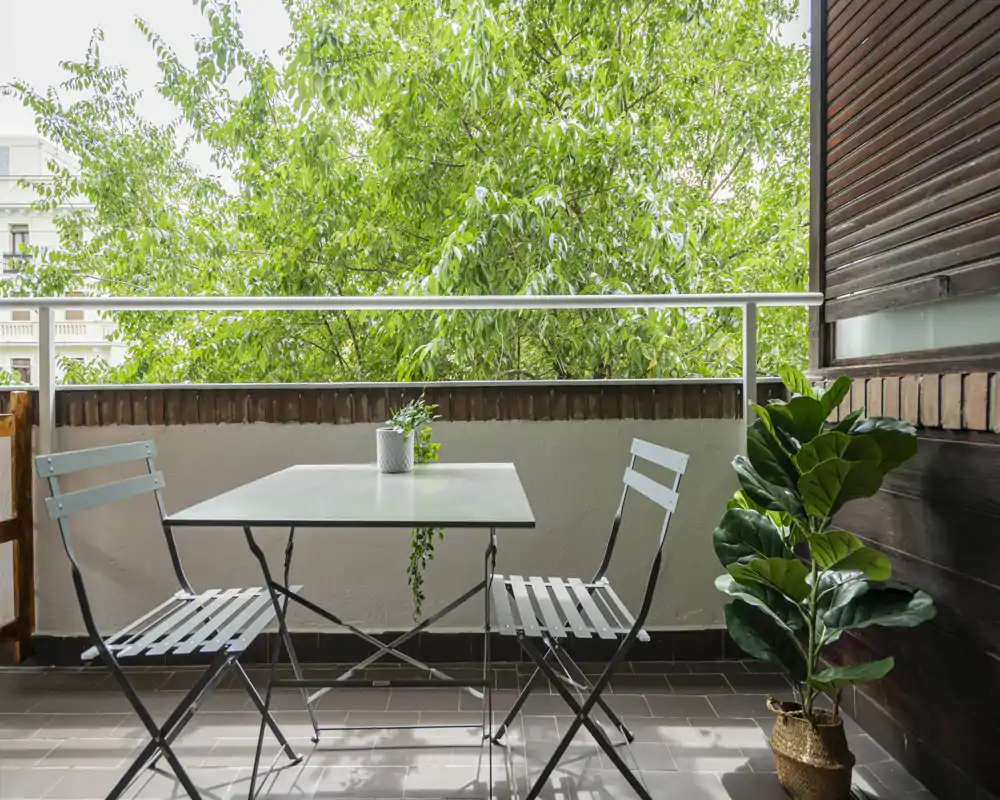 Balcone soleggiato con un piccolo tavolo in metallo e due sedie pieghevoli, decorato con una pianta in vaso e una vista verde — un angolo accogliente per il caffè mattutino.