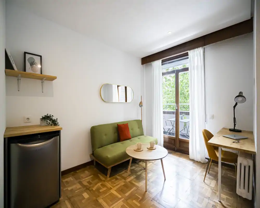 Zona giorno luminosa e curata con un divano verde compatto, tavolino rotondo e scrivania vicino al balcone — perfetta per rilassarsi o lavorare con luce naturale.