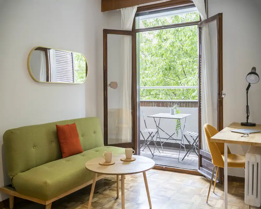 Zona giorno luminosa e accogliente con divano verde, tavolino rotondo in legno e porta finestra aperta su un piccolo balcone con tavolino da bistrò. La luce naturale e gli accenti caldi rendono lo spazio invitante per rilassarsi o lavorare.