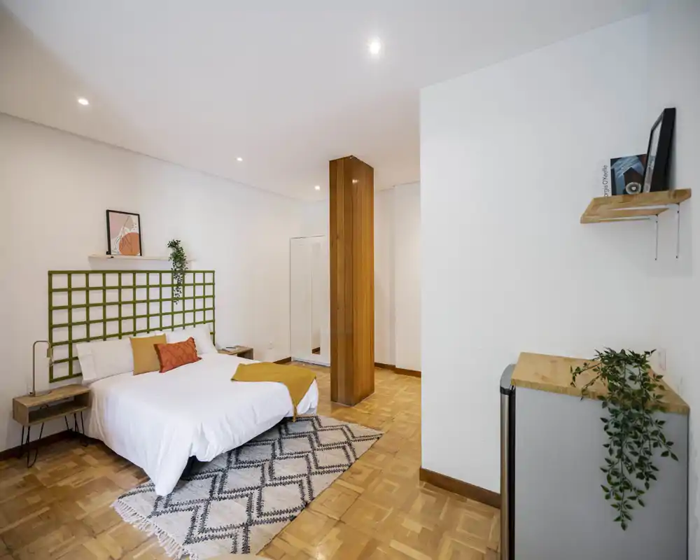 Camera da letto luminosa e moderna con letto matrimoniale, tappeto geometrico e pavimenti in legno; arredo minimale e illuminazione piacevole.