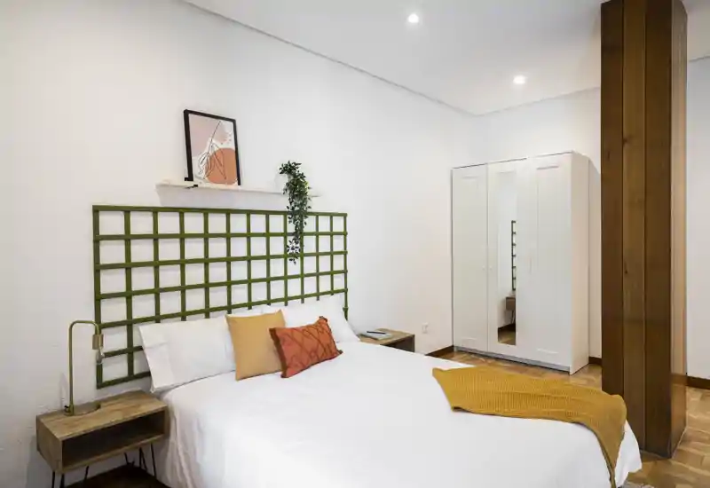 Camera da letto luminosa e moderna con letto matrimoniale, cuscini decorativi e plaid senape; pareti bianche, comodini e un armadio con specchio per pratico spazio di stoccaggio.