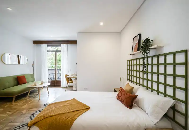 Camera da letto luminosa e di design con letto matrimoniale comodo, tessili caldi e un piccolo angolo salotto vicino al balcone. Grande finestra/porta che offre molta luce naturale e vista sul verde.