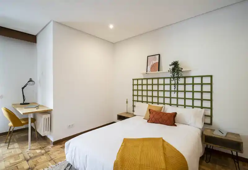 Camera da letto luminosa e stilosa con letto matrimoniale, cuscini decorativi e un plaid senape. Un piccolo scrittoio con sedia è accanto al letto, con decorazioni minimal e una pianta che rendono l'ambiente accogliente.