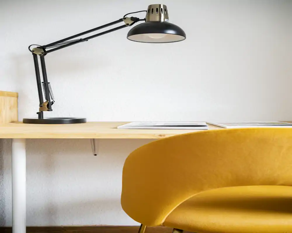 Angolo lavoro accogliente con una lampada da scrivania regolabile e una poltroncina gialla senape accanto a una scrivania in legno minimalista — perfetto per mostrare un piccolo soggiorno o un angolo studio.