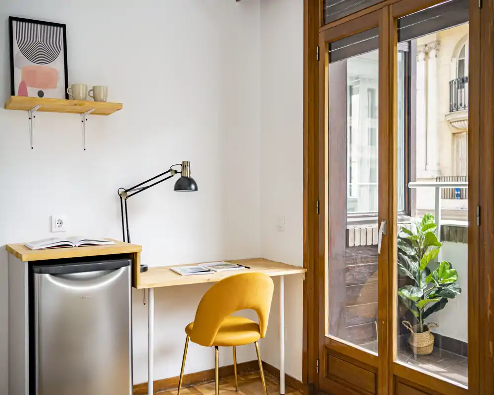 Zona giorno luminosa e compatta con scrivania in legno, sedia gialla moderna e ampie porte a vetro che si affacciano su un piccolo balcone — perfetta come angolo smart working.