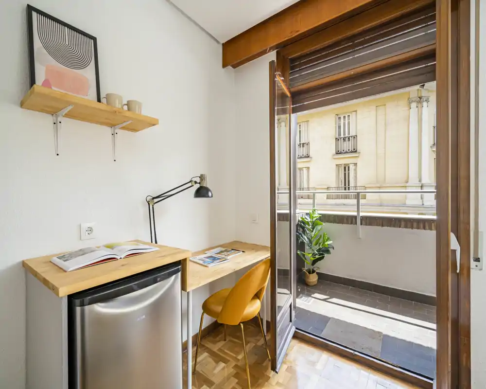 Zona giorno luminosa e compatta con scrivania in legno, sedia moderna e balcone che lascia entrare molta luce naturale — ideale come spazio di lavoro o monolocale.