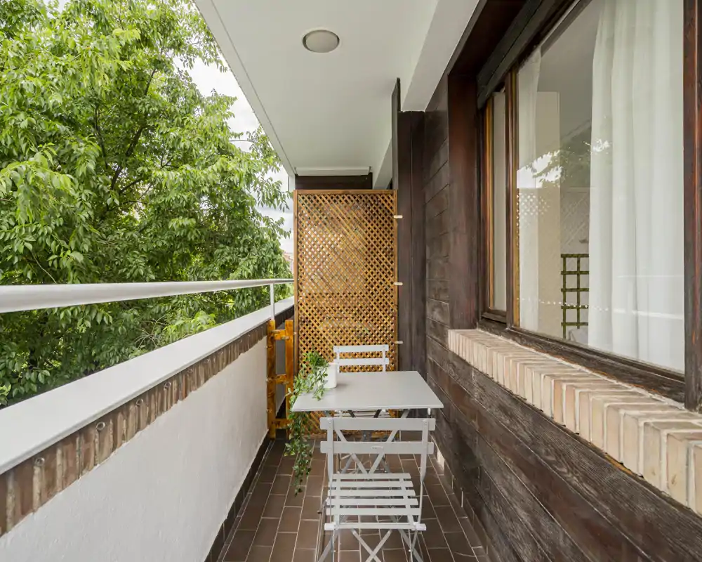 Balcone stretto e soleggiato con un piccolo tavolo e due sedie, parete in legno e alberi verdi all'esterno — perfetto per un caffè mattutino.