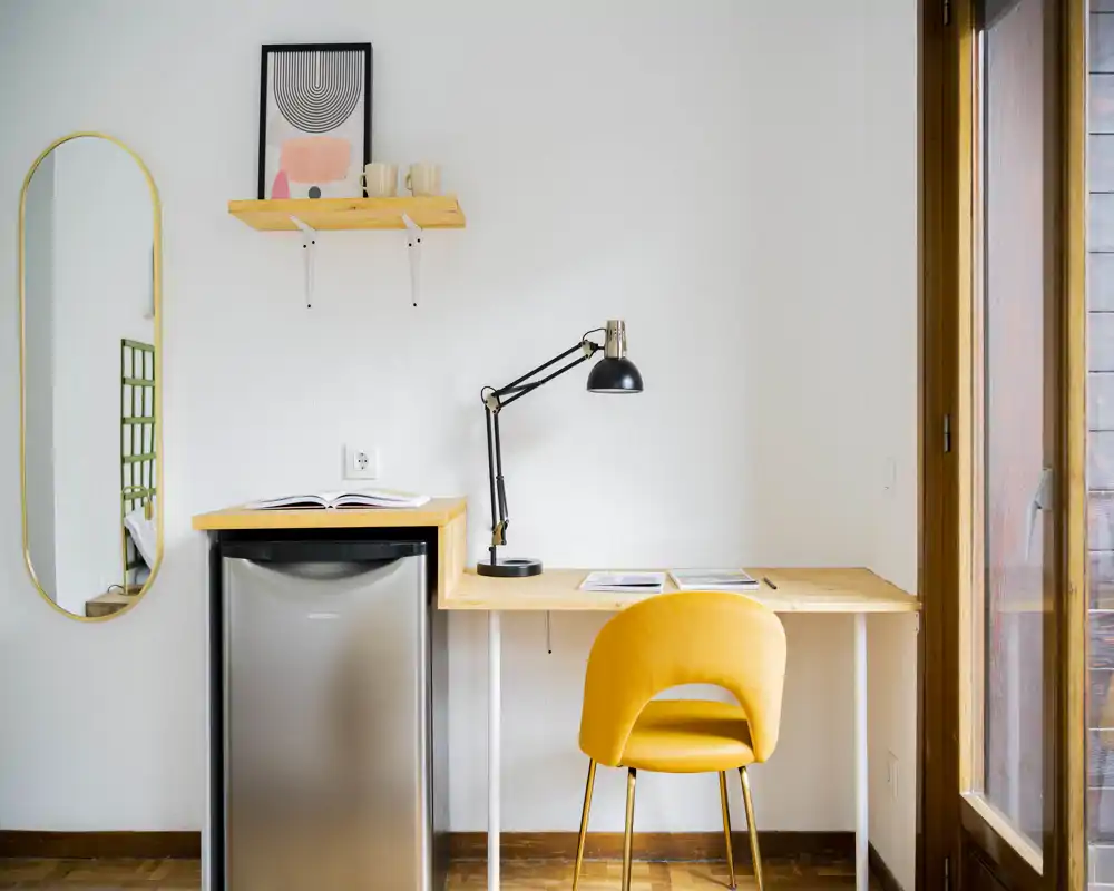 Angolo lavoro luminoso e minimale con scrivania in legno, sedia imbottita gialla, lampada regolabile e un piccolo frigorifero sotto. La luce naturale entra da una grande porta finestra, creando uno spazio di lavoro pulito e funzionale.