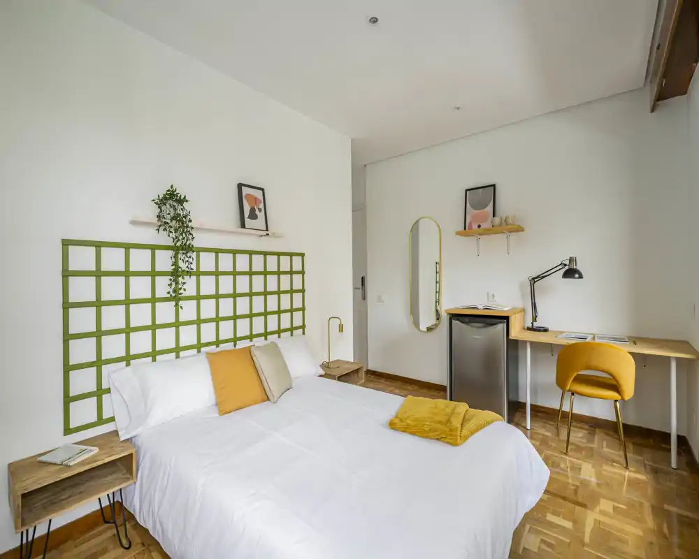Camera da letto luminosa e moderna con letto matrimoniale completamente visibile, accenti gialli caldi e un pratico angolo studio con scrivania e sedia. Biancheria bianca e pavimento in parquet creano un ambiente accogliente.
