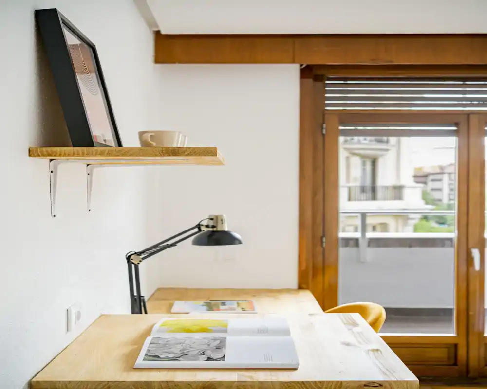Angolo luminoso con scrivania in legno e lampada vicino alla porta del balcone; mensola minimal e una sedia accogliente creano un confortevole angolo living/working con luce naturale.