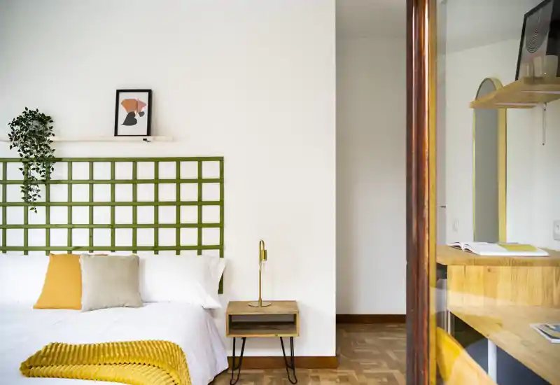 Camera da letto luminosa e curata con testiera a griglia verde, biancheria bianca e accenti gialli caldi come coperta e cuscini.