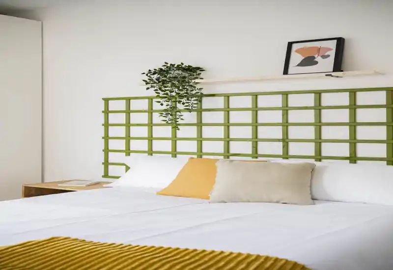 Camera da letto luminosa e curata con ampio letto ben rifatto, cuscini colorati, testiera a griglia verde e una pianta sospesa che aggiunge freschezza.