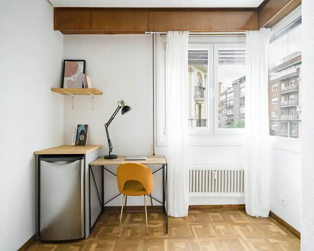 Coin salon lumineux et minimaliste avec un bureau, une lampe de lecture et une chaise jaune près d'une grande fenêtre apportant de la lumière naturelle.