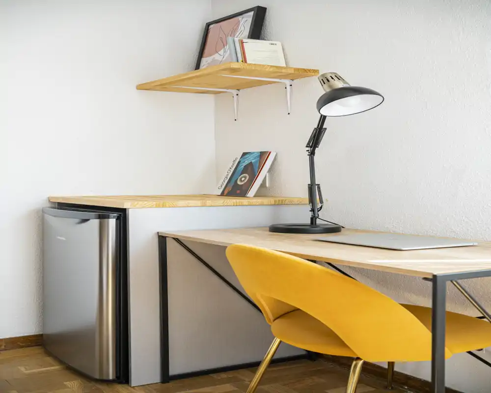 Coin kitchenette compact avec plan de travail en bois, mini-frigo inox et un espace bureau soigné avec lampe moderne et chaise jaune — lumineux et fonctionnel pour les courts séjours.